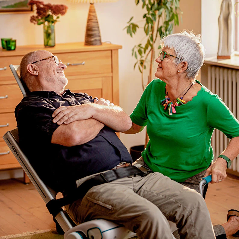 A close-up stock photograph shows a caregiver operating the pump for a Mangar Camel inflatable lifting cushion, which is gently lifting an elderly man from the floor in a home setting.